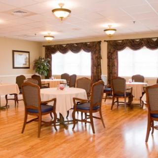 A well-lit dining room with multiple tables and chairs, featuring wooden flooring and large windows with curtains.
