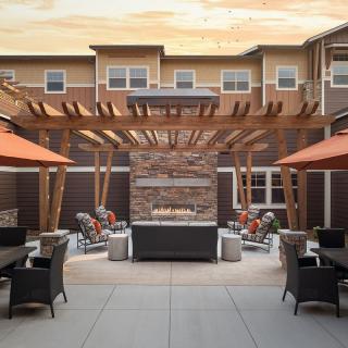 A modern outdoor patio with a pergola and central fireplace surrounded by seating and umbrellas.