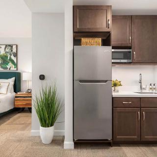 A modern apartment featuring an open kitchen with dark cabinetry and a stainless steel refrigerator, adjacent to a bedroom area with stylish decor.