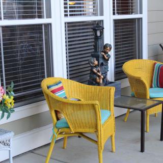 A cozy patio setup with yellow wicker chairs and colorful cushions, accompanied by a decorative flower arrangement beside a window.