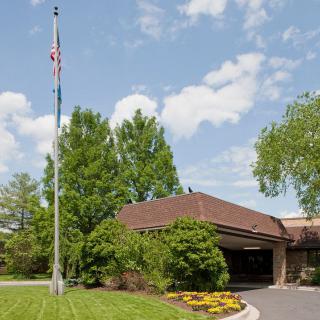 The image shows a building with a flagpole in front, surrounded by trees and landscaping under a partly cloudy sky.