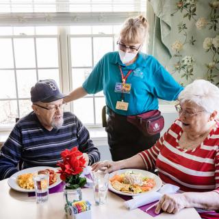 A caregiver assists two elderly individuals during a meal in a nursing home setting.