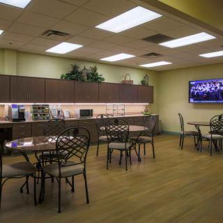 A spacious and well-lit break room featuring tables, chairs, a kitchenette, and a wall-mounted television.