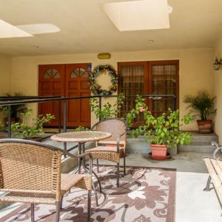 A cozy patio with wicker chairs and a table surrounded by potted plants, featuring wooden doors adorned with a decorative wreath under warm sunlight.
