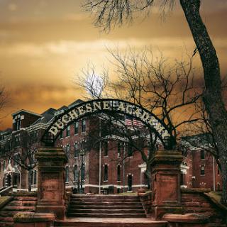 The iconic entrance of Duquesne Academy, framed by barren trees under a dramatic sunset.