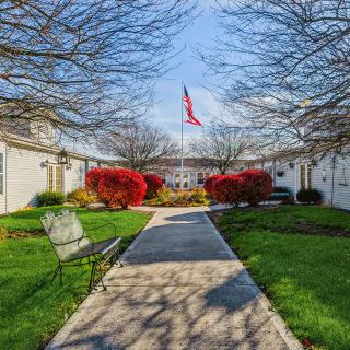 A scenic outdoor pathway leads to a building, flanked by benches and vibrant red bushes, with a flag waving in the background.