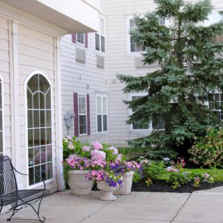 A peaceful patio area with a bench, potted flowers, and lush greenery next to a building.