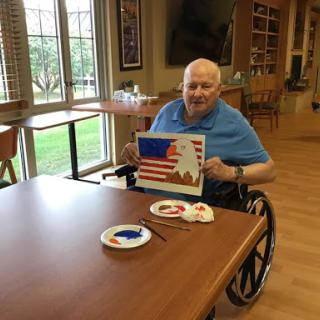 An elderly man proudly showcases his painting of the American flag during an art activity.