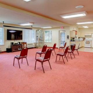 An empty community room with red chairs arranged in rows, a large television, and a kitchen area in the background.