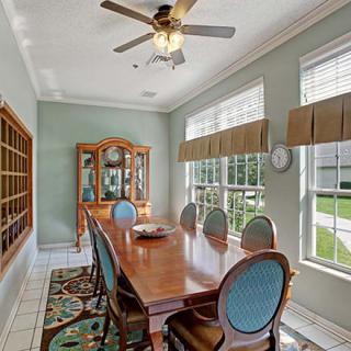 A well-lit dining room featuring a long wooden table with matching chairs, large windows with curtains, and a decorative cabinet.