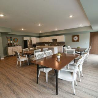 A spacious dining area with multiple tables and chairs, featuring a modern kitchen in the background and wood flooring.