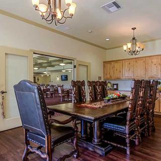 A spacious dining room with a large wooden table and ornate chairs, featuring chandeliers and a view into the kitchen area.