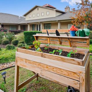 A backyard garden with a raised wooden planter box and gardening tools in front of a house.