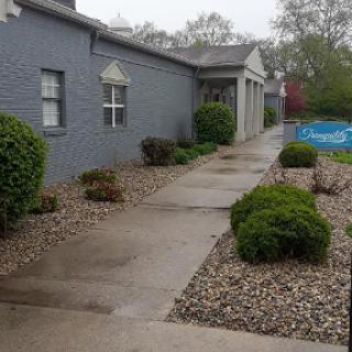 A smooth pathway lined with bushes and gravel, leading to a building during rainy weather.