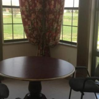 A round table with chairs by a large window with floral curtains, overlooking a green landscape.