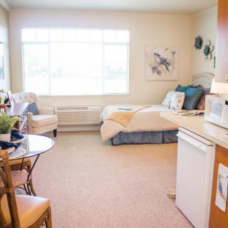 A cozy studio apartment with ample natural light and minimalist decor, featuring a bed, chair, and small kitchenette area.