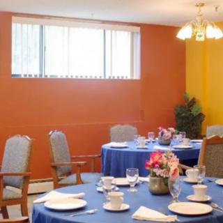 A dining room with tables set with blue tablecloths and a floral centerpiece, surrounded by chairs against an orange wall.