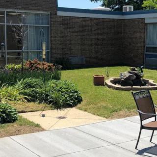 A peaceful garden patio area with a chair, surrounded by greenery and a building.