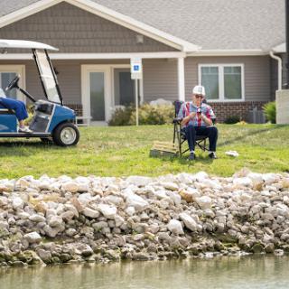 A man relaxes by a pond, enjoying the outdoors next to his golf cart in a residential area.