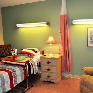 A hospital-style bedroom with a single bed, nightstand, lamp, and chair.