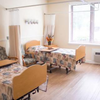 A bright healthcare room with two beds, a curtain divider, and flowers on the table.