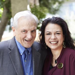A smiling elderly man and a woman posing together outdoors.