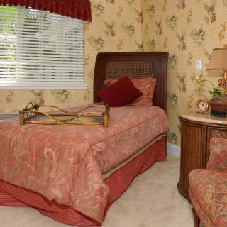 A vintage-style bedroom featuring a single bed with floral bedding, decorative pillows, and ornate wooden furniture against a backdrop of floral wallpaper.