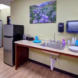 A small office kitchen area with a sink, countertop, refrigerator, and wall art.