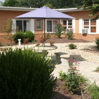 A peaceful garden patio with a large umbrella providing shade over a picnic table, surrounded by plants and a stone pathway.