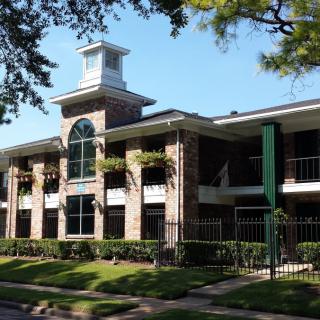 A two-story residential apartment building with a distinctive arched window in the center and surrounded by trees and a gated entrance.