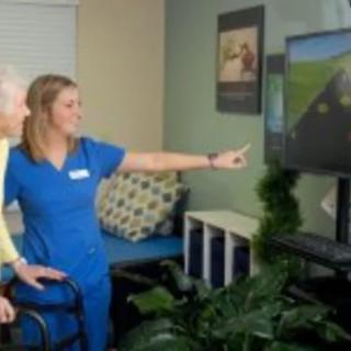 A healthcare professional assists an elderly patient with physical therapy using a video screen.