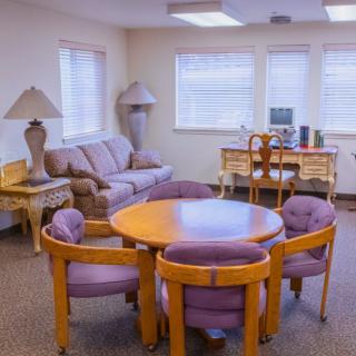 A cozy living room with a round wooden table surrounded by chairs, a sofa, and a desk near the windows.