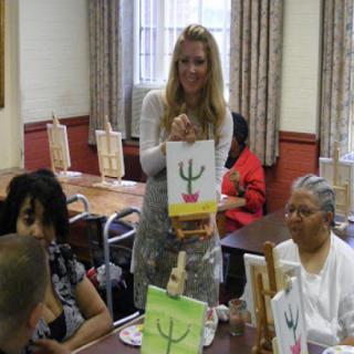 A group of people participate in an indoor art workshop, with one person holding a painting.