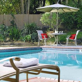 A tranquil outdoor patio area featuring a swimming pool, surrounded by greenery and set with a table and chairs under an umbrella.
