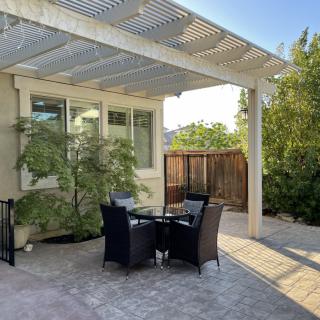 A serene patio area with a pergola providing shade over a set of outdoor table and chairs, surrounded by lush greenery.