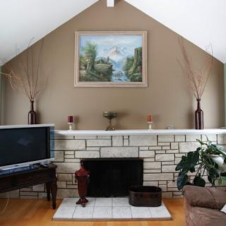 A cozy living room with a central fireplace, adorned with a landscape painting and flanked by decorative vases. A television sits to the side, and an indoor plant adds a touch of greenery.