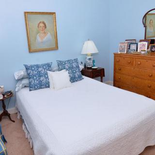 A cozy bedroom with blue walls, featuring a neatly made bed adorned with decorative pillows, a bedside lamp, a dresser with a mirror, and a portrait on the wall.