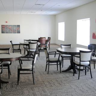 An empty meeting room with tables and chairs arranged on a carpeted floor, lit by natural light from large windows.