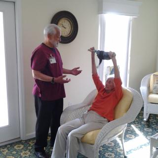 A caregiver interacts with a senior who is exercising indoors.