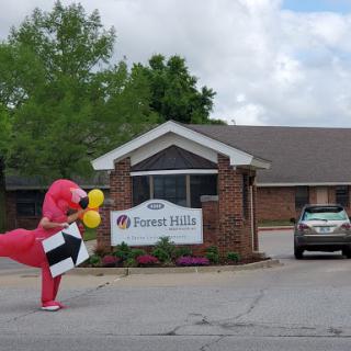 A person in a dinosaur costume holding balloons and a sign outside a building.