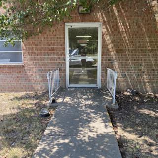 The entrance of a brick building with a clear pathway and shadows cast by surrounding trees.