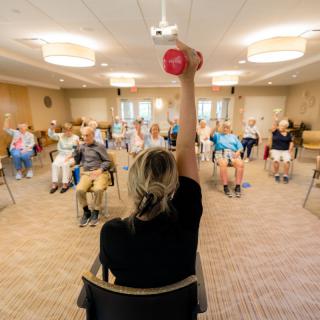 A group of seniors participating in a seated exercise class led by an instructor holding a dumbbell.