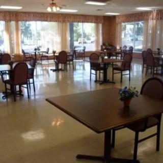 An empty dining room with tables and chairs arranged neatly, large windows with curtains allowing natural light.