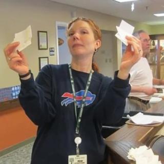 A woman indoors holding up pieces of paper with a confident expression.