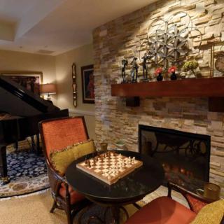 A cozy living room with a chess set on a table, featuring a fireplace with a stone wall and a grand piano in the background.