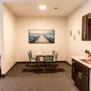 A modern dining area featuring a table with four chairs, artwork on the wall, and a small kitchenette.