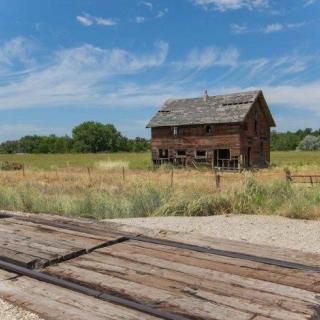 An abandoned wooden house stands in a rural landscape under a clear blue sky.