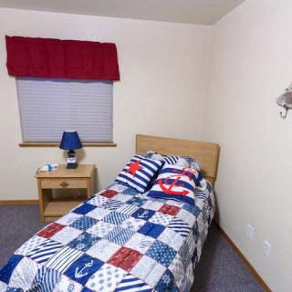A small bedroom featuring a single bed with nautical-themed bedding, a nightstand with a blue lamp, and a red curtain.