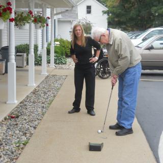 An elderly man plays mini golf with a woman assisting him.