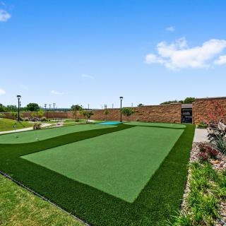 A landscaped outdoor area with artificial turf, surrounded by greenery and a clear blue sky.